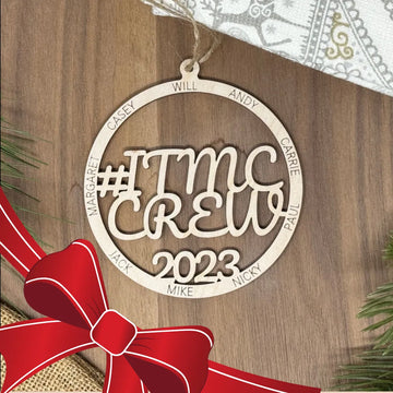 Decorative ornament with '#ITMC CREW 2023' text on a wooden background with a red bow.