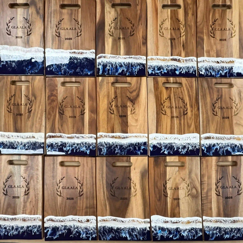 A batch of wooden serving boards with blue ocean-wave resin and matching engraved event designs.