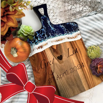 Wooden cutting board with engraved names and date, surrounded by decorative items on a textured surface.